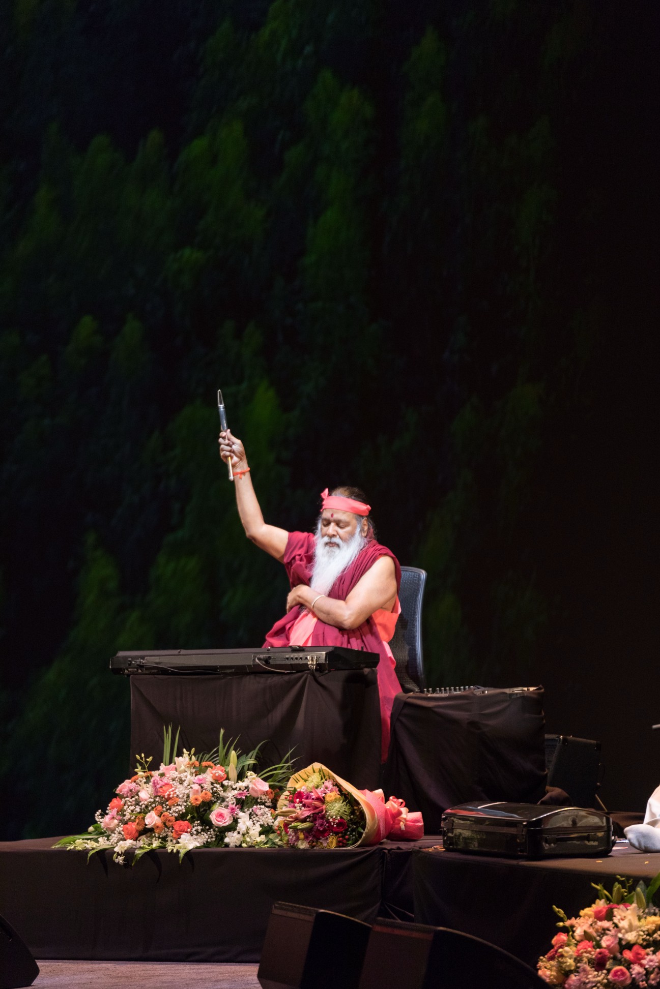 Sri Swamiji raising arm on stage — forest backdrop